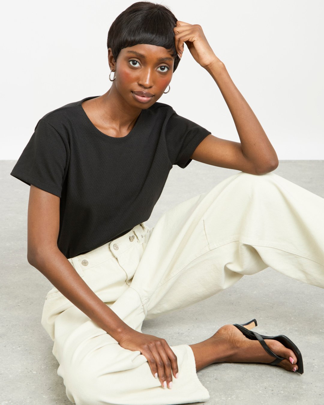 Women wearing ivory denim jeans with a black crew neck t-shirt sitting on the floor wearing toe-post heeled sandals.